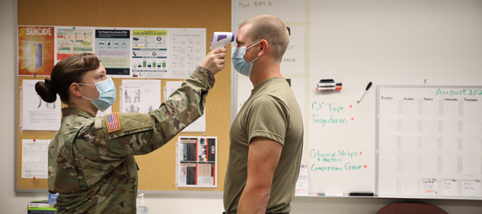 Photo of a woman (wearing a medical mask and army fatigues with a USA flag on the shoulder) taking the temperature of a man (also wearing a medical mask and an army green t-shirt) using a forehead thermometer. The background is an office with a bulletin board and dry erase board.
